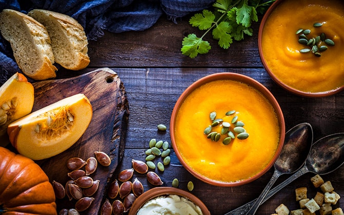 A top-down photo of two bowls of pumpkin soup topped with pumpkin seeds, surrounded by fall slices of rustic bread, a pair of spoons, a smattering of croutons, a sprig of cilantro, and sliced and whole pumpkin