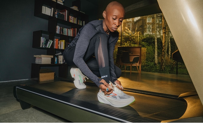 Cynthia Erivo ties her shoe on a treadmill, with a bookshelf and natural setting in the background
