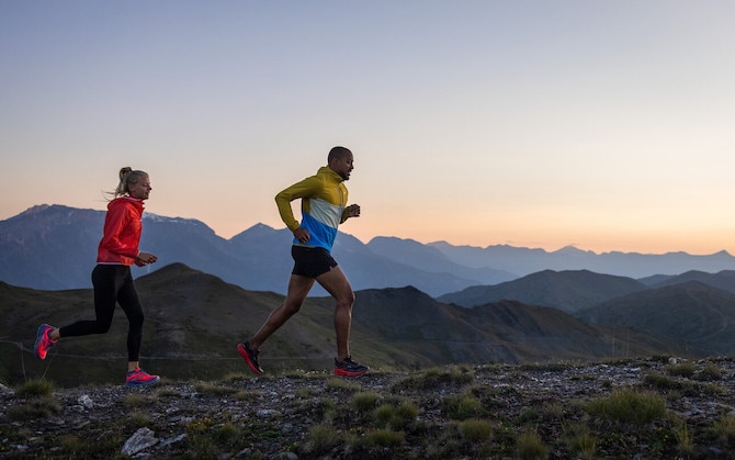 Dos corredores trotan por la cresta de una montaña con un amanecer o una puesta de sol detrás.
