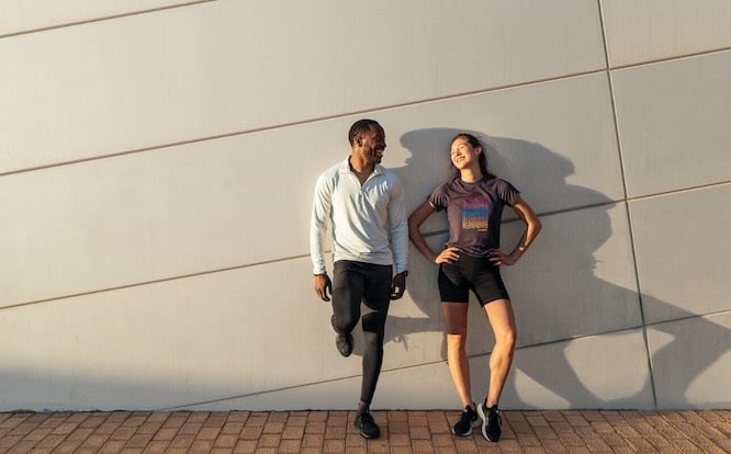 Two smiling runners lean against a wall after a run.