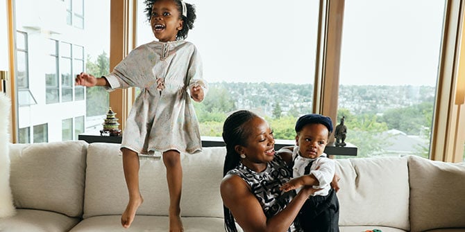 Olu holding one of her kids while the other jumps on the couch