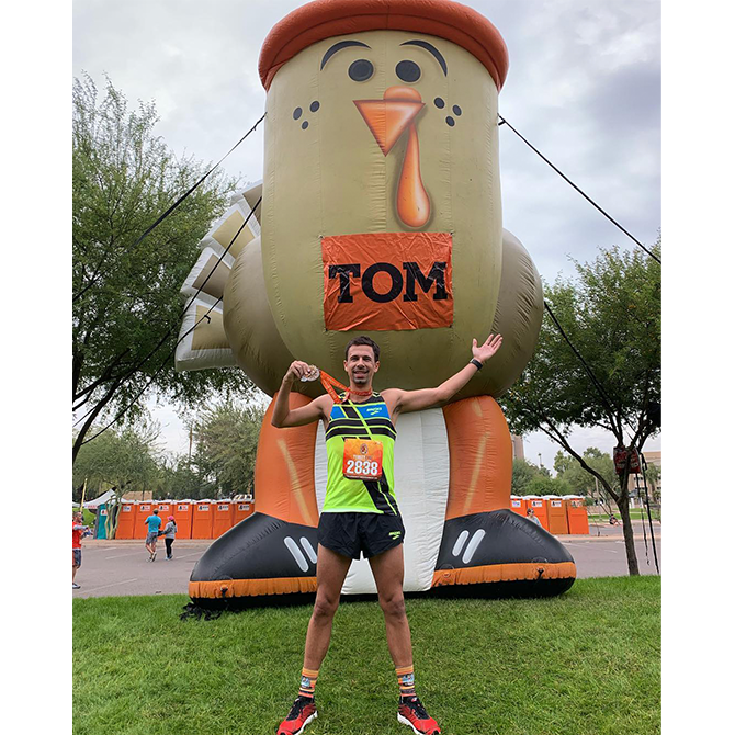 A male runner poses with a medal in front of a giant inflatable turkey that is wearing a sign that says “Tom.”