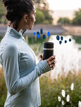 A woman holding a coffee tumbler looks out at a lake.