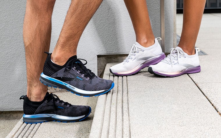 Two runners stand on concrete steps, one wearing black and blue Ricochet shoes, one wearing white and purple Ricochet shoes.
