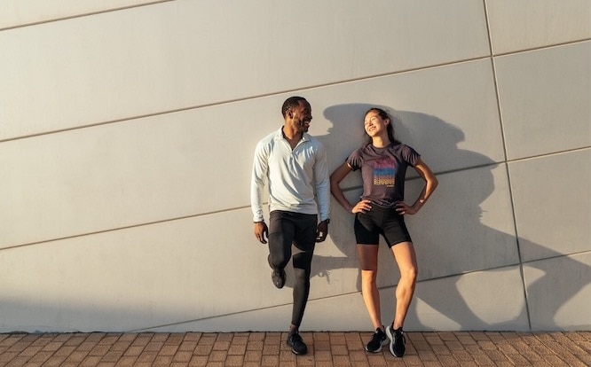 Two smiling runners lean against a wall after a run.