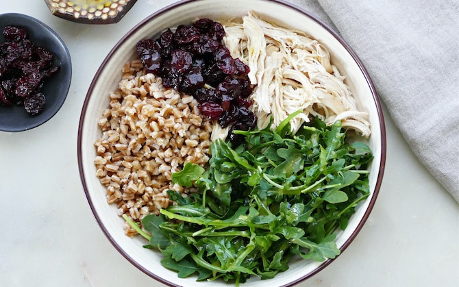 Een bovenaanzicht van een farro-bowl met versnipperde kip, rucola en gedroogde kersen.
