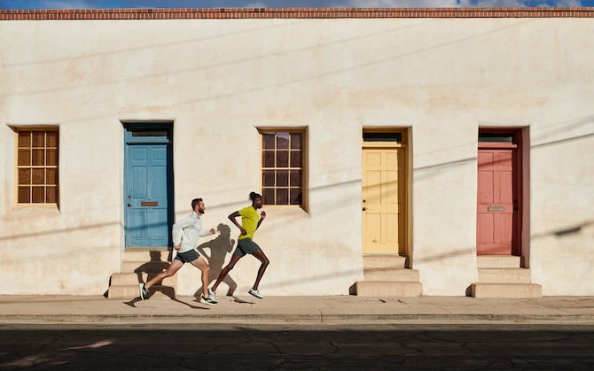 Two men running on the sidewalk in front of a building with coloured doors