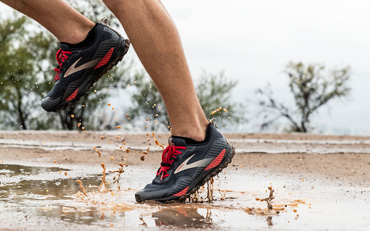 Gros plan sur les pieds d’un coureur traversant des flaques de boue avec les Cascadia GTX.
