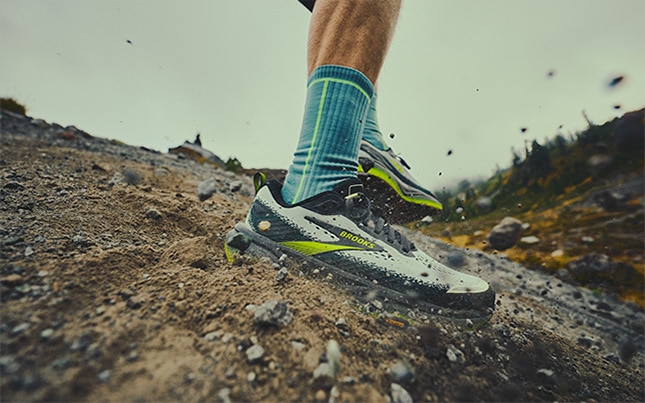 Close-up of someone running in the Cascadia 20 trail running shoes on a gravel trail in the mountains with rocks and dirt