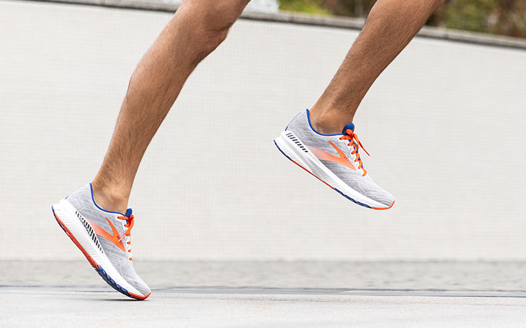 Gros plan sur les jambes d’un coureur en mi-foulée portant des chaussures Ravenna grises et orange devant un mur gris.