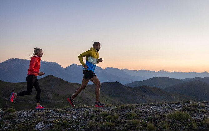Two runners jog along a mountain ridge with a sunrise or sunset behind them.