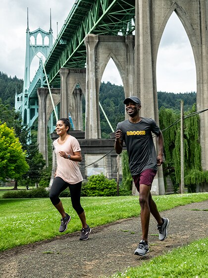 Two people run on a grass-line trail with a big bridge in the background.