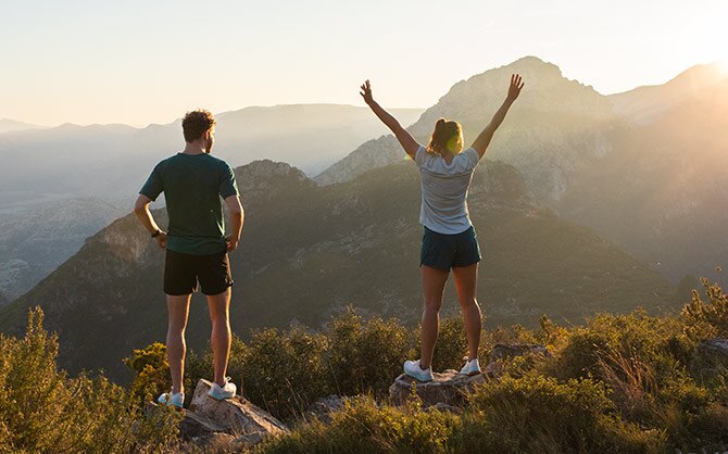 Two runners, one with hands on his hips and the other with hands raised high in celebration, look out at a picturesque mountain landscape. 