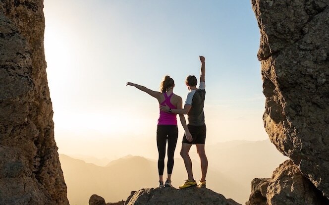 Two runners hold up their arms to celebrate a successful trail run while standing on a rock.