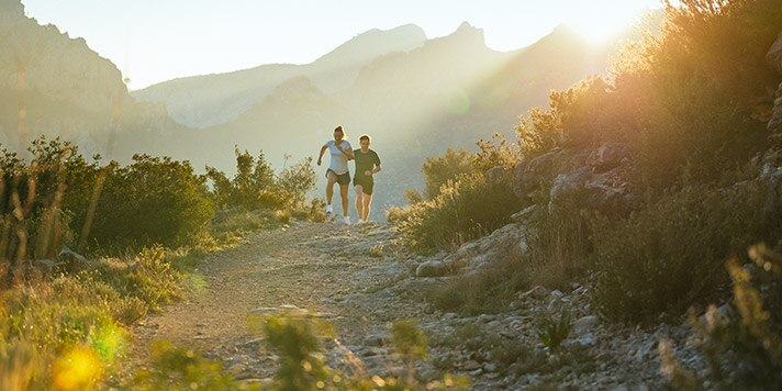 Two runners ascend a mountain trail, with hills behind them.