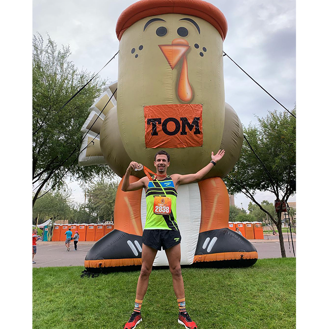 A male runner poses with a medal in front of a giant inflatable turkey that is wearing a sign that says “Tom.” 