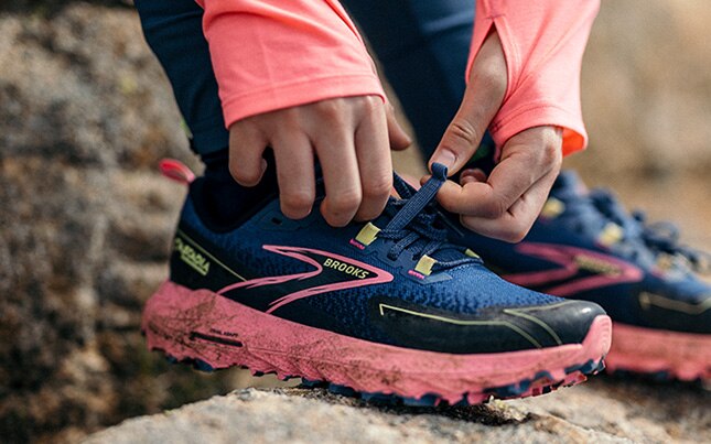 Gros plan d’une personne qui attache ses chaussures de course en sentier Cascadia 18, avec de la terre autour du bas des chaussures et un terrain rocheux en arrière-plan