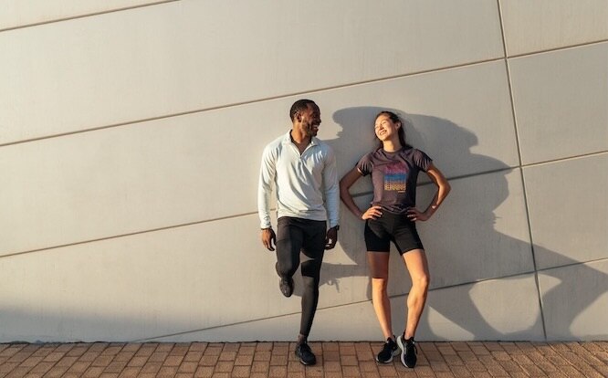 Two smiling runners lean against a wall after a run.