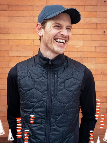 A smiling man wearing a hat, vest, and long-sleeve shirt poses in front of a brick background. 