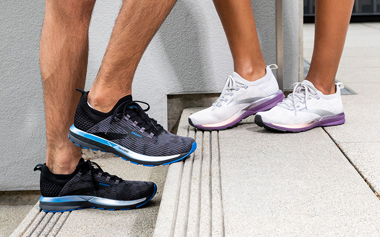 Two runners stand on concrete steps, one wearing black and blue Ricochet shoes, one wearing white and purple Ricochet shoes.