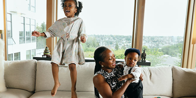 Olu holding one of her kids while the other jumps on the couch
