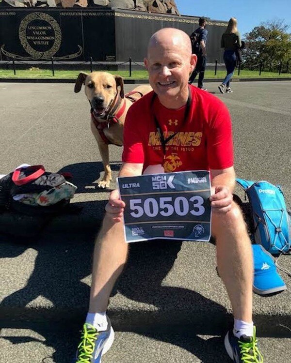 marathon finisher sitting with sign and dog behind him