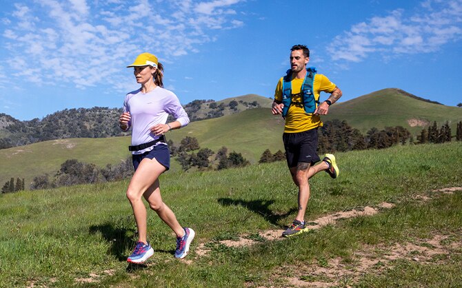Zwei Läufer*innen auf einem Trail mit Aussicht auf grüne Hügel.