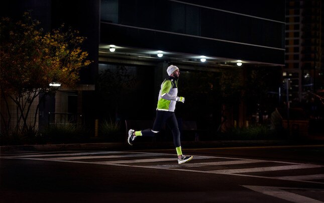 Vista lateral de un corredor corriendo de noche por la carretera con equipo reflectante.