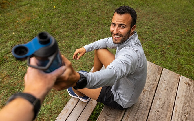 coureur en phase de récupération, à qui un autre coureur tend une gourde. Il est assis sur une marche et porte un haut de sport gris à manches longues, un short de running noir et une paire de chaussures Brooks Glycerin GTS 21 bleu marine.