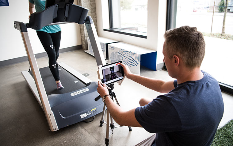 A man uses a tablet to observe a runner who is running on a treadmill in a lab. 