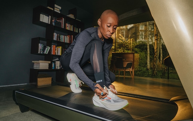 Cynthia Erivo ties her shoe on a treadmill, with a bookshelf and natural setting in the background