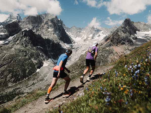 Two people wearing Brooks gear walk along a grassy trail, a blue sky and mountain in the background