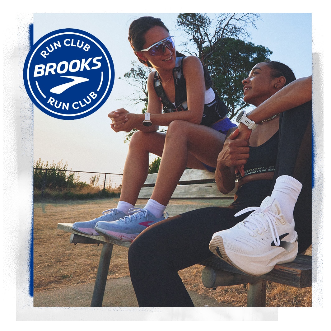 Two women wearing Brooks gear sitting on a bench and talking on a sunny day
