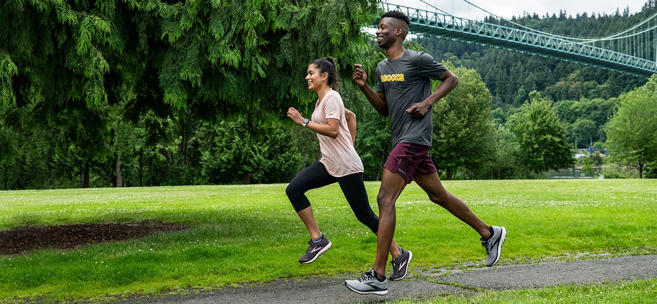 Female runner in a light pink running short, black tights and Brooks Running shoes and a male runner in a black shirt, maroon shorts and Brooks Running shoes running together in a park full of trees and grass with a blue bridge in the background. 