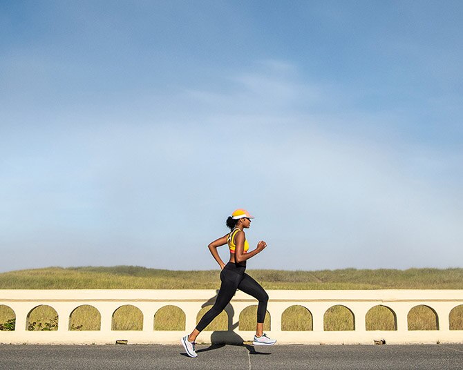 A photo of a woman wearing a yellow Drive Mesh Run Bra running by an open field.