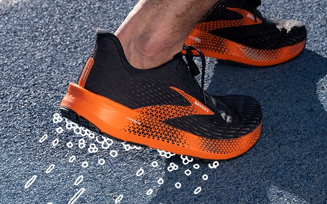 A close-up image of a foot mid toe strike wearing a black and orange Hyperion Tempo. Small illustrated bubbles are along the sole of the shoe . Longer illustrated bubble trail away from the sole of the shoe.