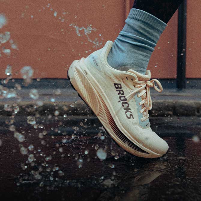 A person wearing Brooks shoes runs outside, water from a puddle splashing behind them