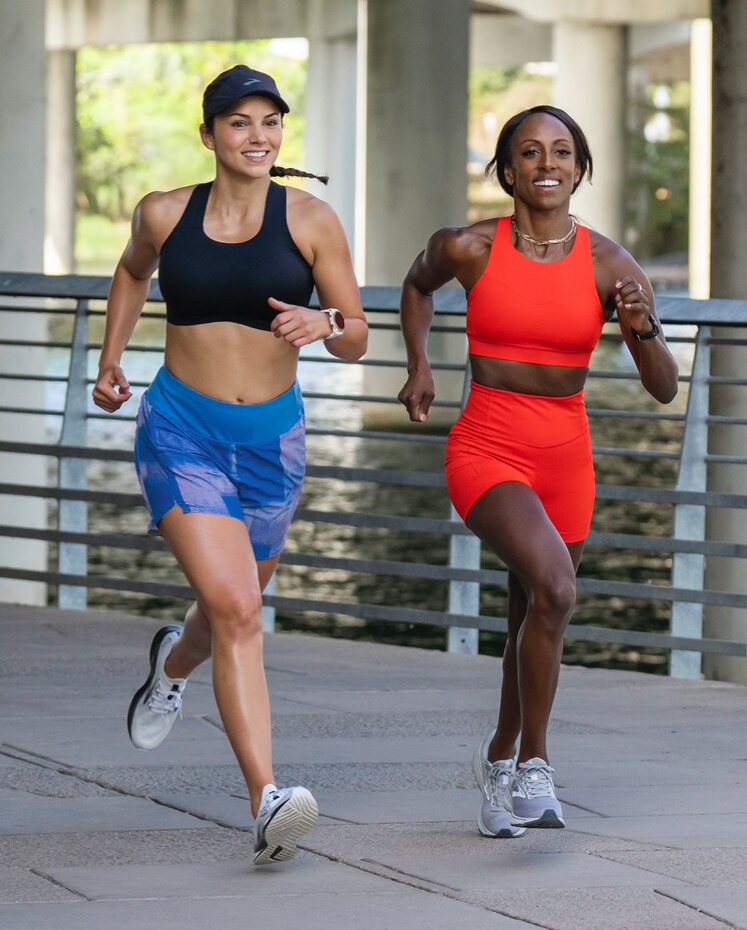 Deux femmes courent dans la rue, une portant une brassière course à dos nageur Dare bleue et l’autre portant une brassière de course noire Dare à dos dégagé.
