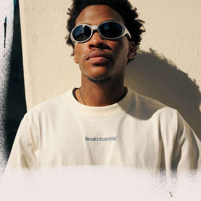 A man wearing a Brooks Running shirt looks ahead, leaning against a white wall
