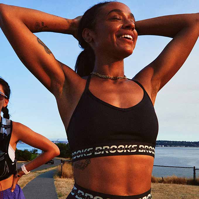 A woman wearing the PR Elite Bra stretches outside, a smile on her face